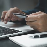 Photo showing just a person’s hands in front of a laptop computer, with a credit card in hand, as well as a note pad and pen next to them.