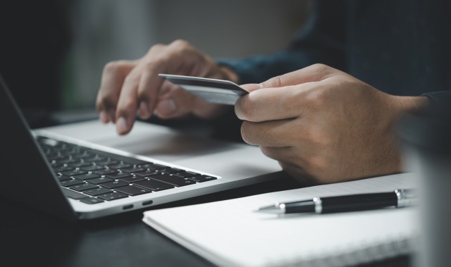Photo showing just a person’s hands in front of a laptop computer, with a credit card in hand, as well as a note pad and pen next to them.