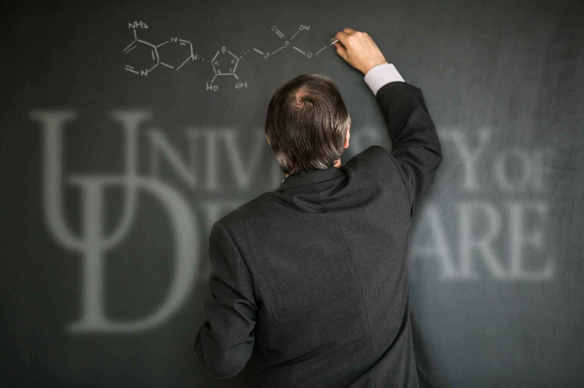 A professor drawing a molecular structure on a chalkboard with the words "University of Delaware" displayed faintly below the molecule.