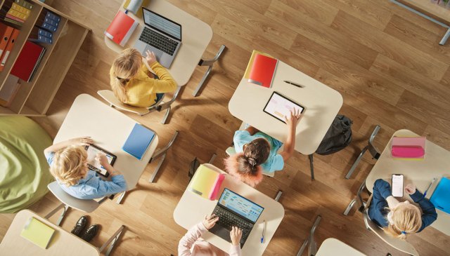 Overhead shot of young children sitting at desks during school and working on laptops and tablets.