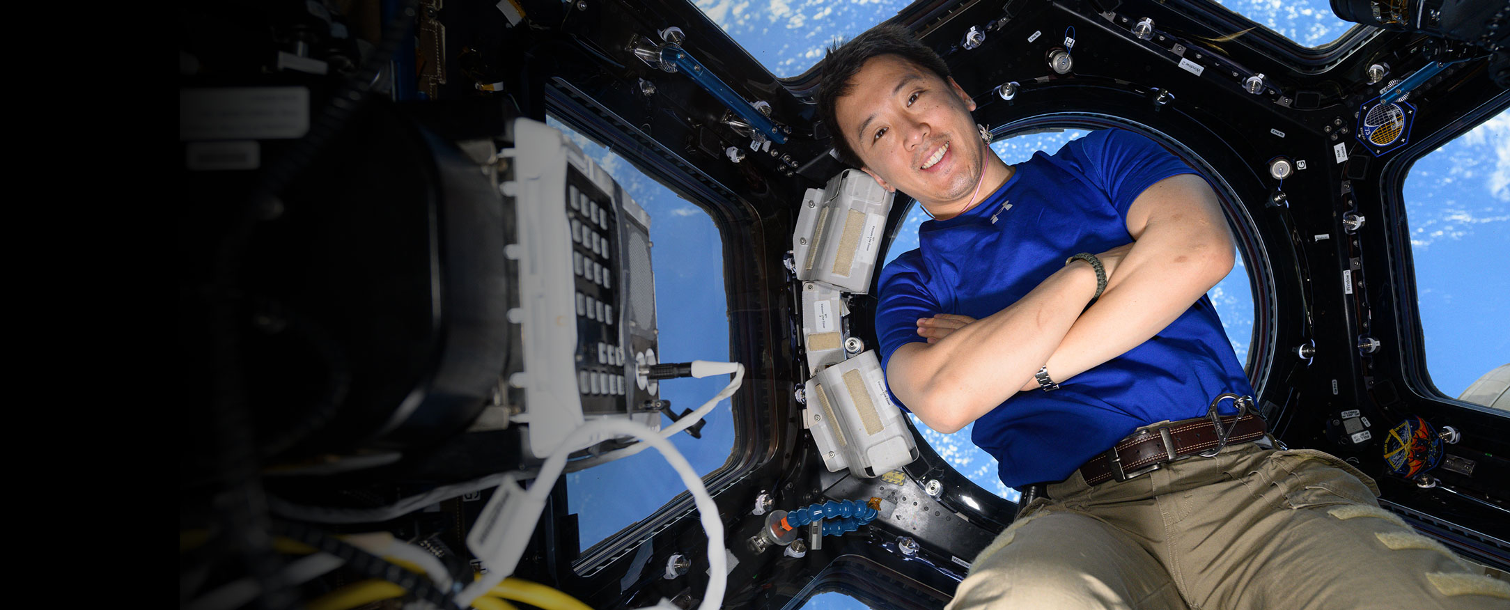 An astronaut with his arms crossed inside the International Space Station. He is leaning up against a large circular window in the cupola, with the Earth behind him.