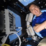 An astronaut with his arms crossed inside the International Space Station. He is leaning up against a large circular window in the cupola, with the Earth behind him.
