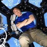An astronaut with his arms crossed inside the International Space Station. He is leaning up against a large circular window in the cupola, with the Earth behind him.