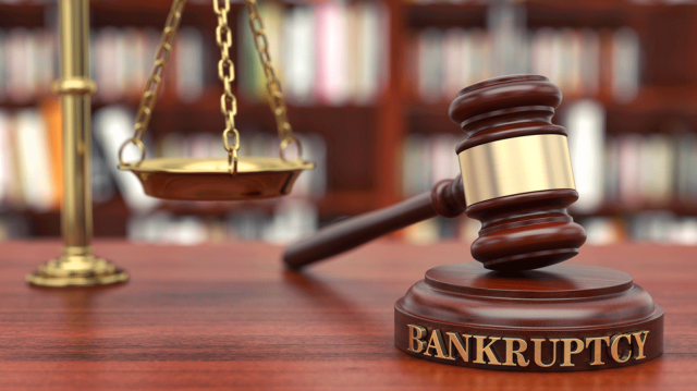 A judge's gavel resting on a gavel block that says "bankruptcy," with the scales of justice and a book case in the background.