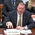 Robert Steinau speaking in front of a mic at a table with audience members behind him