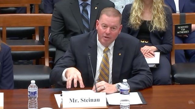 Robert Steinau speaking in front of a mic at a table with audience members behind him