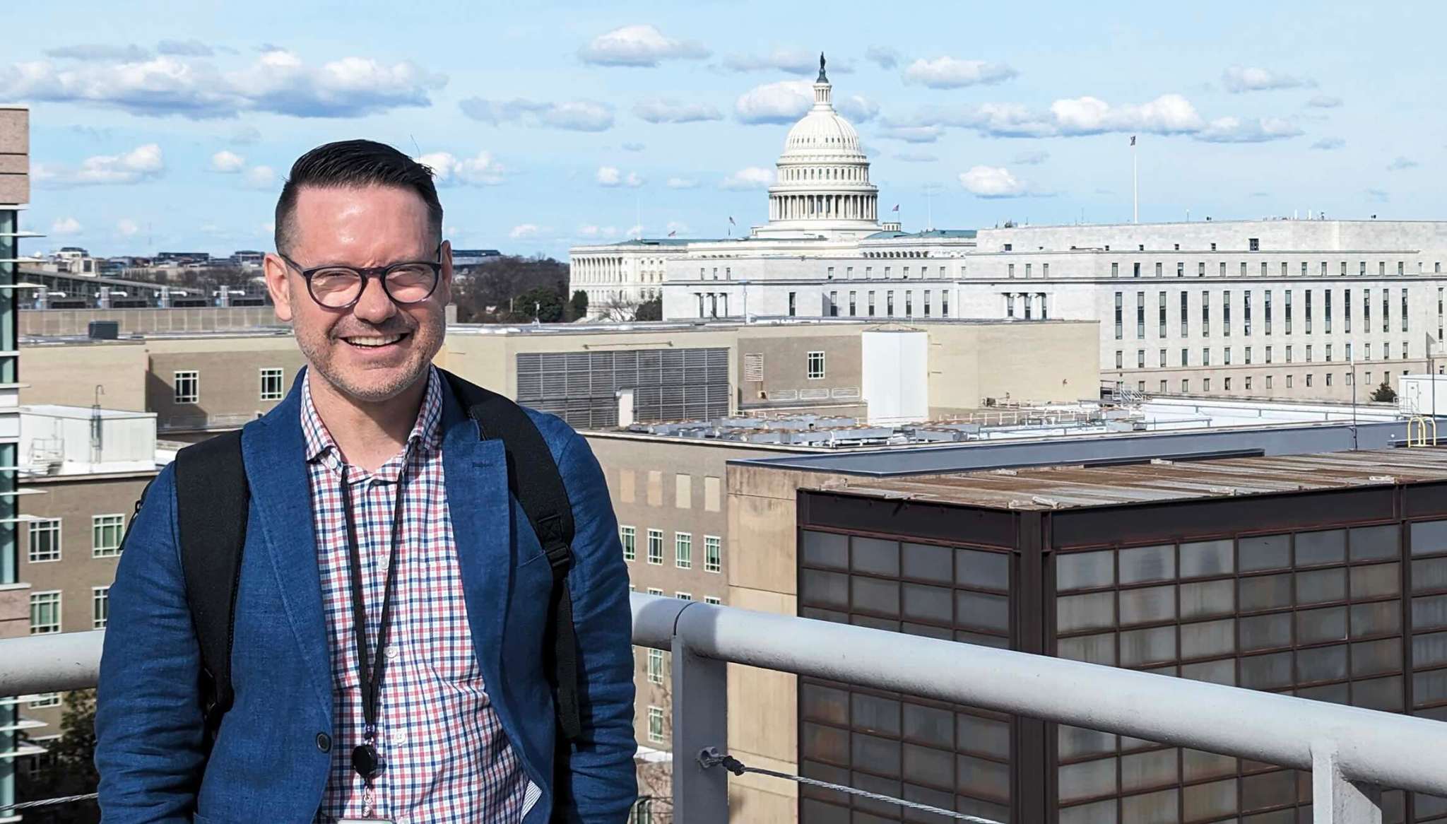 AJ Thibert smiling on the top of a building with a view of the U.S. Capitol in the background.