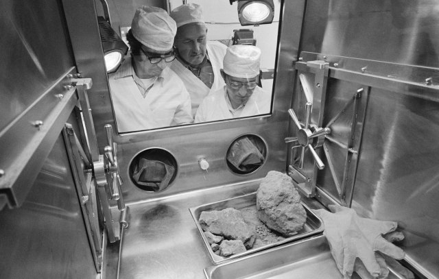 Black and white image of three men in lab coats looking through a window into a room with several Moon rocks in a tray.