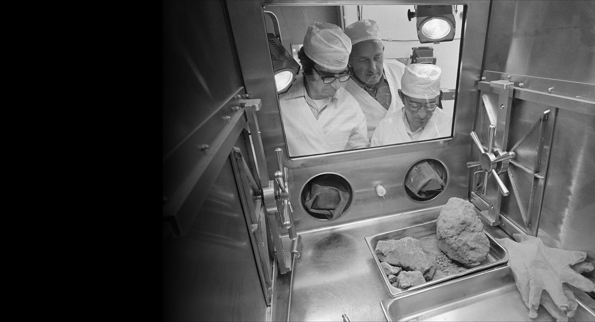 Black and white image of three men in lab coats looking through a window into a room with several Moon rocks in a tray.